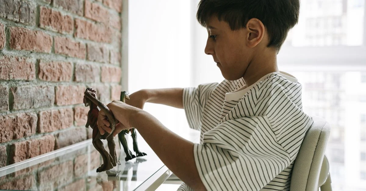 Un niño pequeño jugando con figuras de dinosaurios frente a una pared de ladrillos en una habitación bien iluminada.