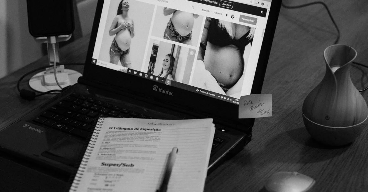 Foto en blanco y negro de un portátil mostrando imágenes de embarazo con un bloc de notas y un bolígrafo al lado.