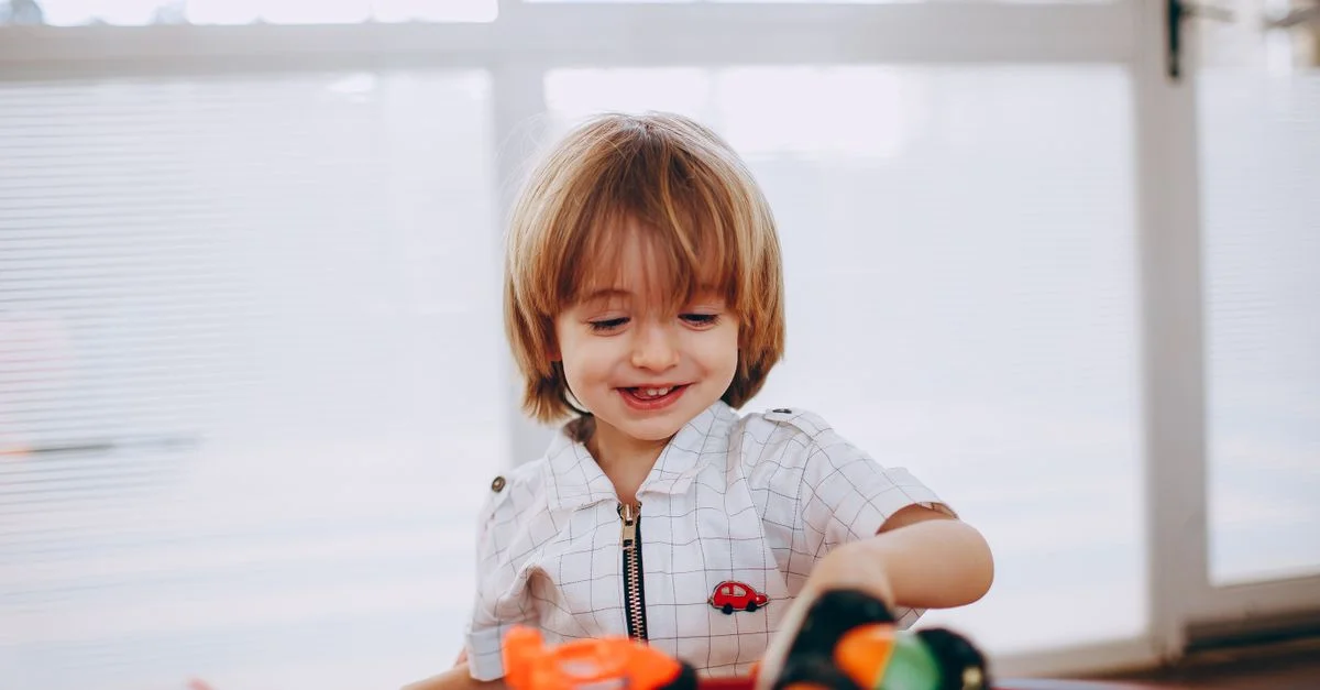 Niño alegre jugando con coches de juguete junto a la ventana, mostrando felicidad y creatividad.