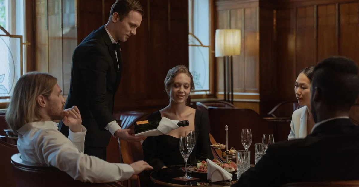 Un camarero sirve vino a un grupo diverso que disfruta de una elegante comida en interiores.