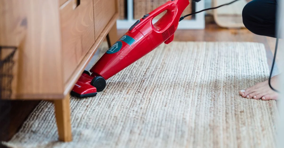 Vista lateral de una persona utilizando una aspiradora roja debajo de los muebles sobre una alfombra para una limpieza eficaz.