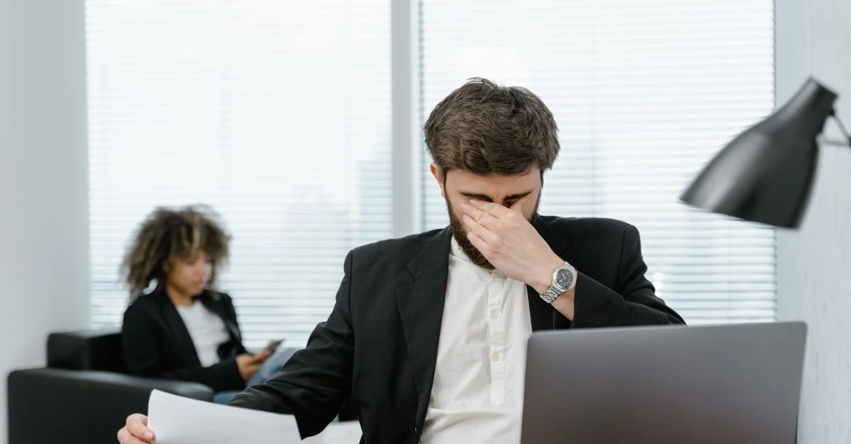 Empresario en una oficina cubriéndose los ojos mientras trabaja en un portátil, sintiéndose estresado.