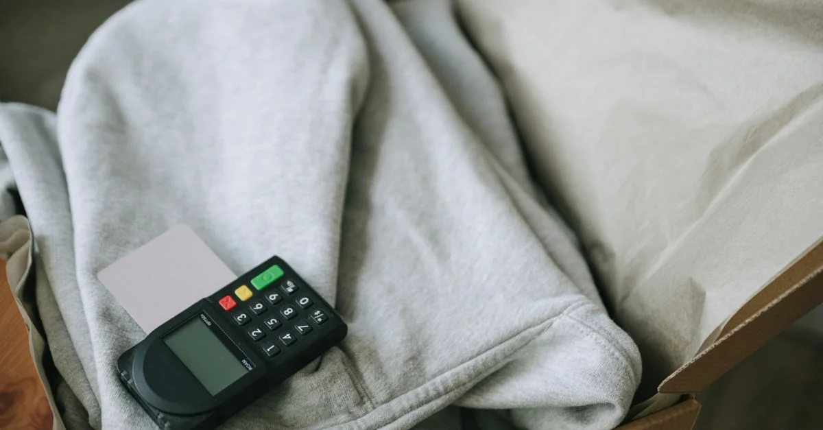 Un terminal de pago y una tarjeta colocados sobre ropa doblada dentro de una caja de envío, indicando empaquetado para comercio electrónico.