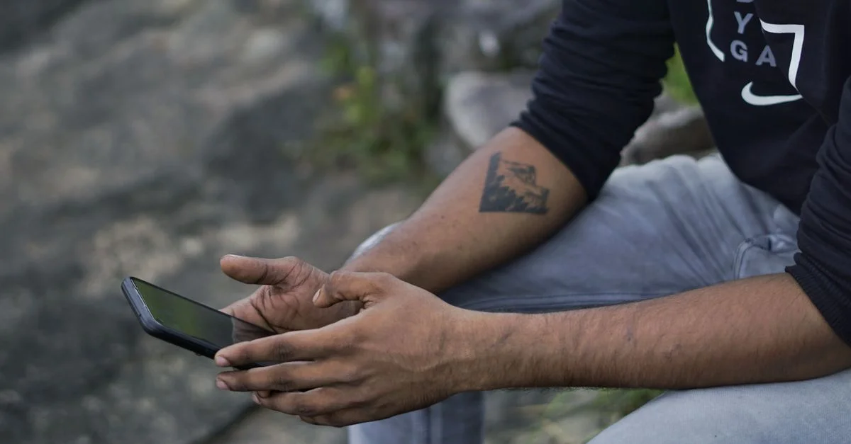 Primer plano de un hombre con un tatuaje en el brazo usando un smartphone mientras está sentado afuera.