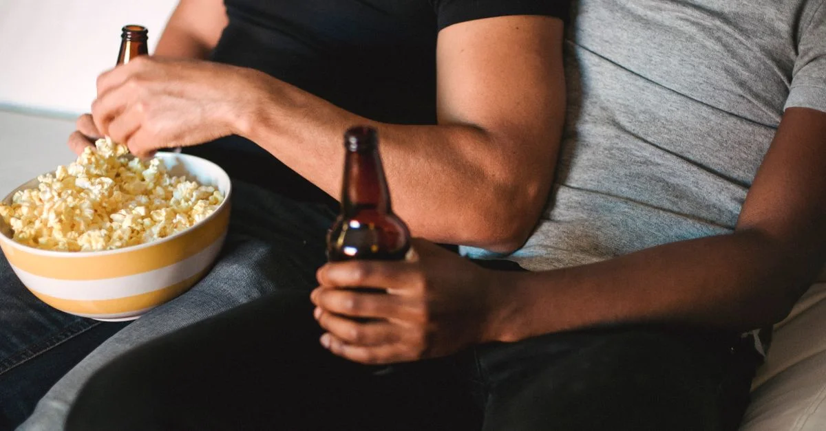 Dos personas disfrutando de un momento relajante con cerveza y palomitas en un sofá acogedor.