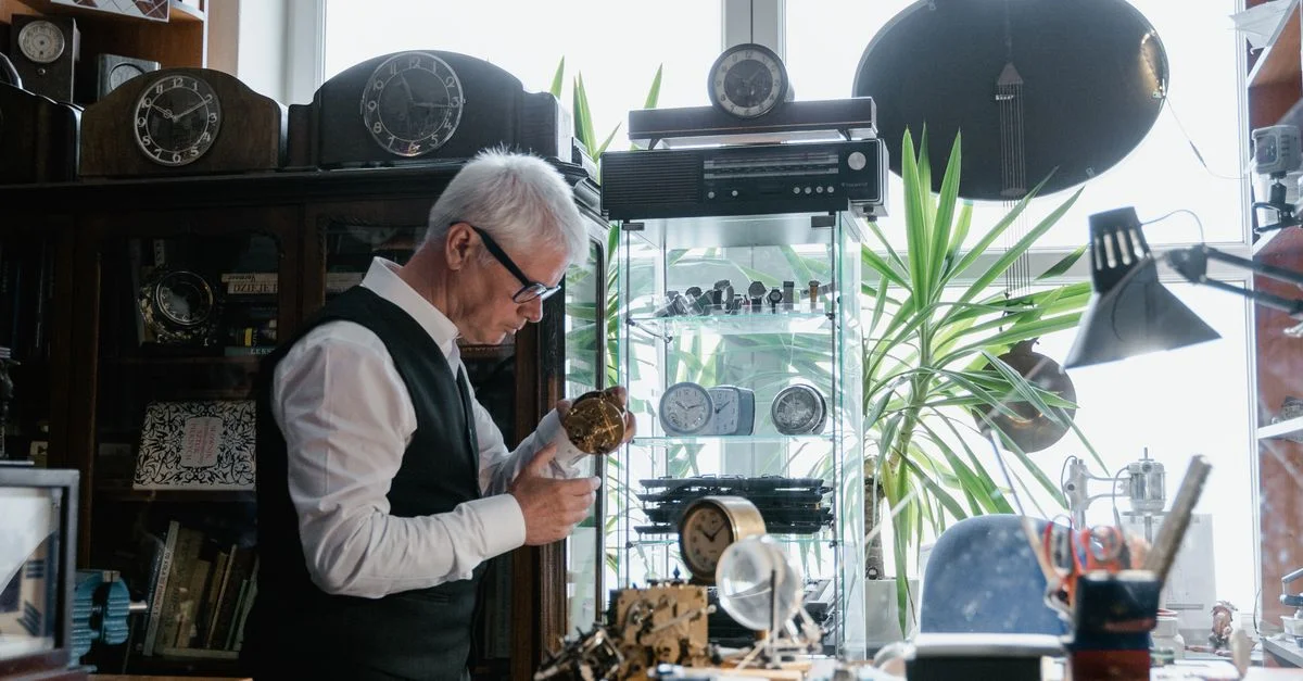 Hombre mayor reparando relojes en un taller rodeado de piezas de tiempo y herramientas.