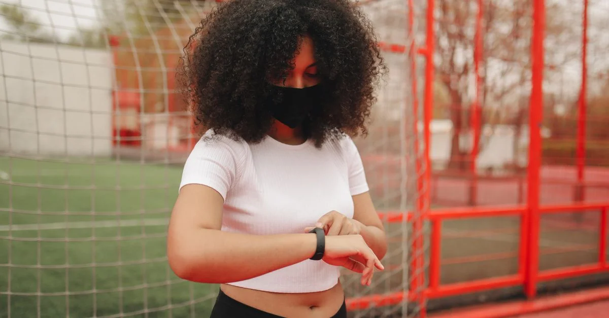 Joven con cabello rizado y mascarilla facial revisando la hora en su reloj de pulsera al aire libre.