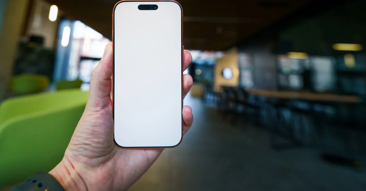 Mano sosteniendo un smartphone con pantalla en blanco, perfecto para fines de maqueta en un entorno de oficina moderno.