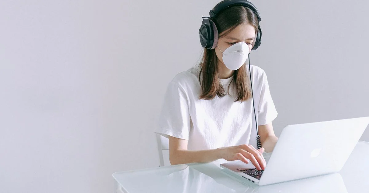 Mujer con mascarilla y auriculares trabajando en un portátil en casa, representando el trabajo remoto durante la pandemia.