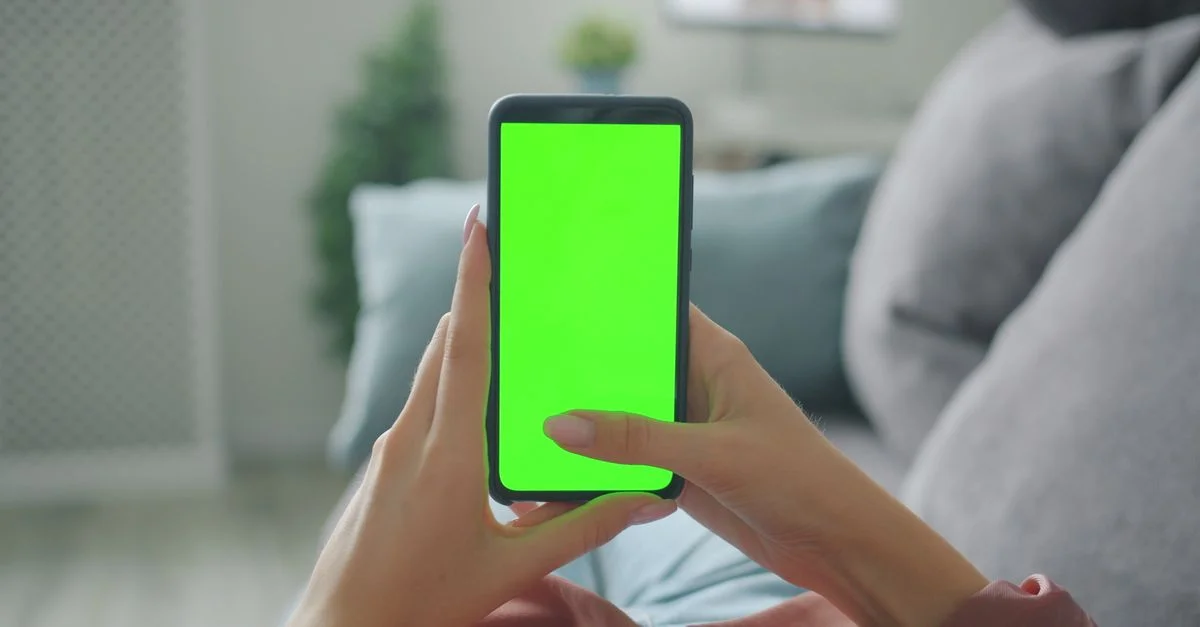 Persona sentada en un sofá en el interior de una casa, sosteniendo un teléfono inteligente con una pantalla verde en blanco.