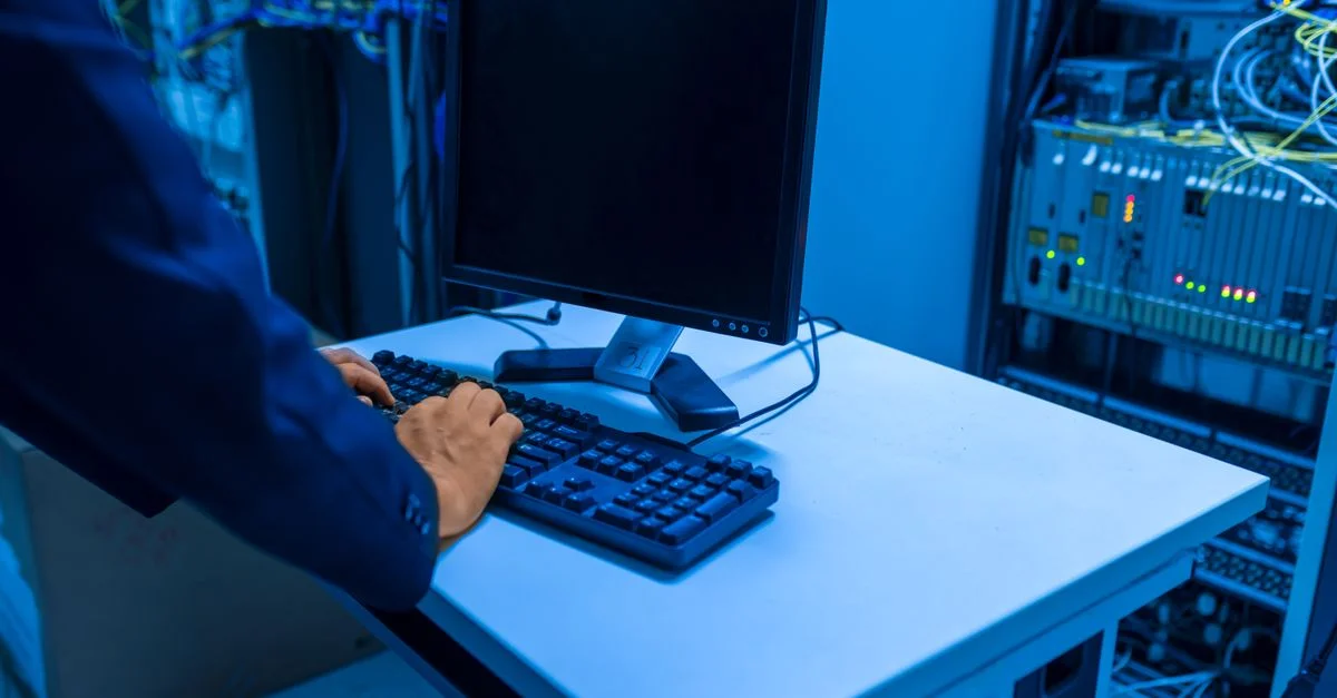 Un profesional de TI opera una computadora en una sala de servidores, gestionando sistemas de red y dispositivos conectados.