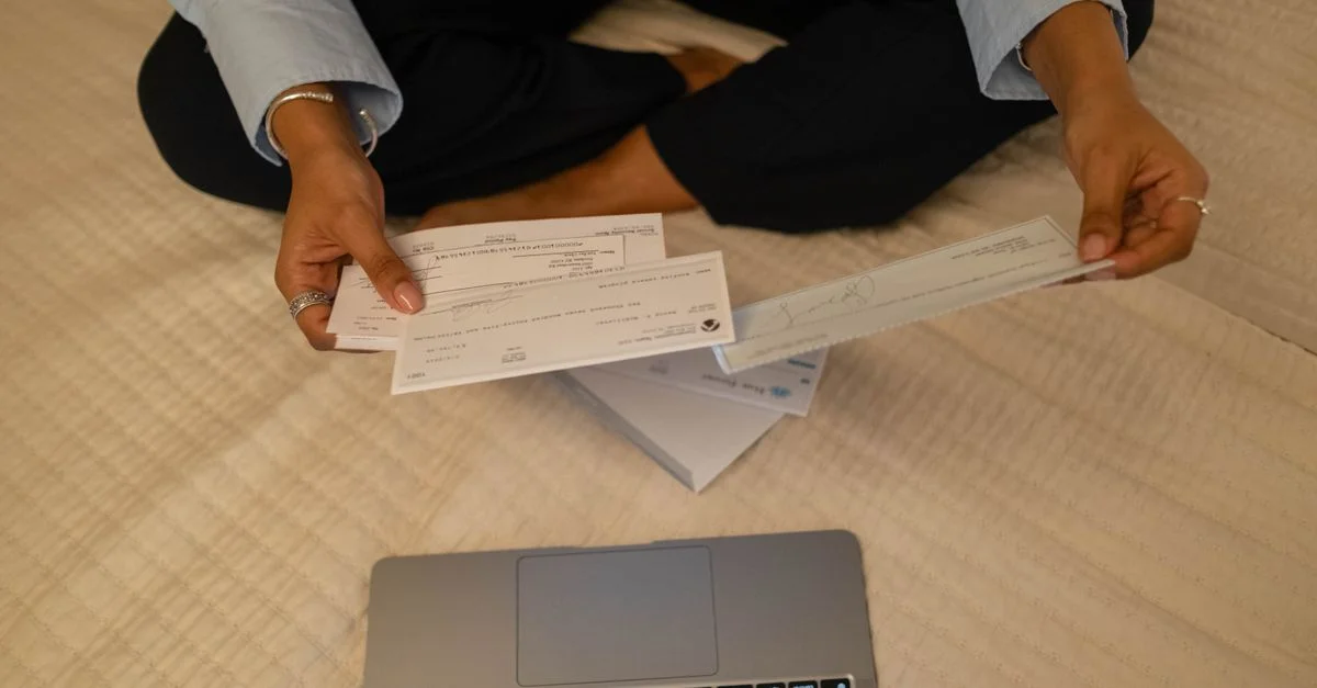 Primer plano de manos sosteniendo cheques junto a un portátil en interiores para tareas financieras.