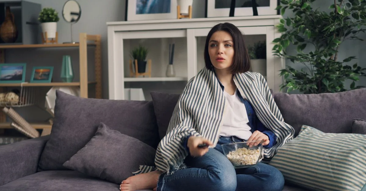 Mujer relajándose con palomitas en un sofá elegante, control remoto en mano.