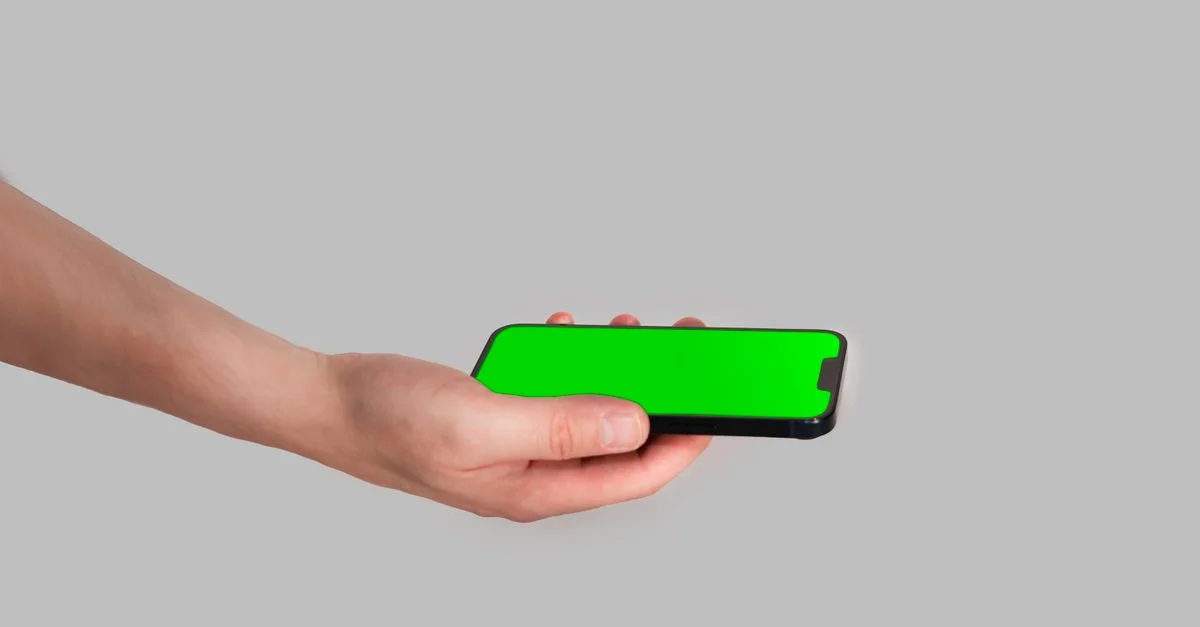 Una mano sosteniendo un teléfono inteligente con una pantalla verde vívida sobre un fondo gris, perfecto para uso en maquetas.