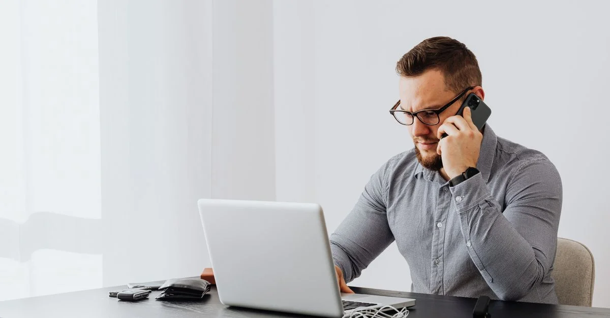 Hombre de negocios concentrado utilizando un smartphone y un portátil en un entorno de oficina, realizando múltiples tareas.