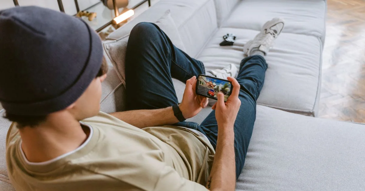 Un joven con ropa casual jugando a un juego móvil mientras se relaja en un sofá en interiores.
