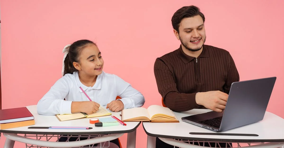 Un maestro guía a un estudiante en un aula con libros y un portátil sobre un fondo rosa.