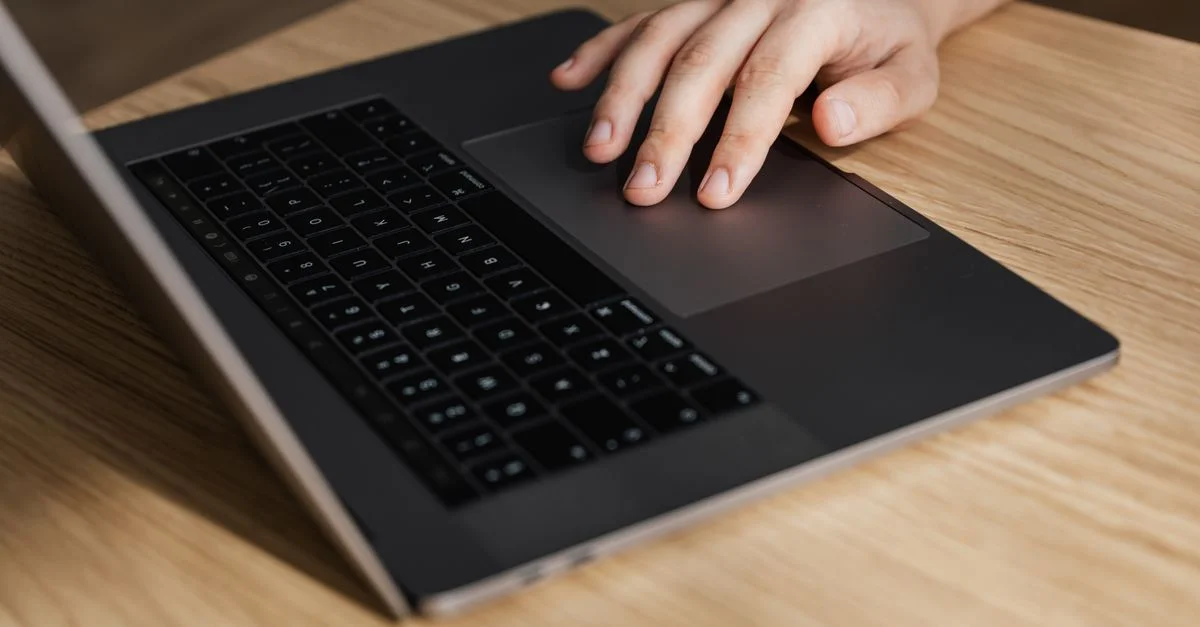 La mano de una persona utiliza el touchpad de un portátil sobre un escritorio de madera, indicando actividad en el espacio de trabajo digital.