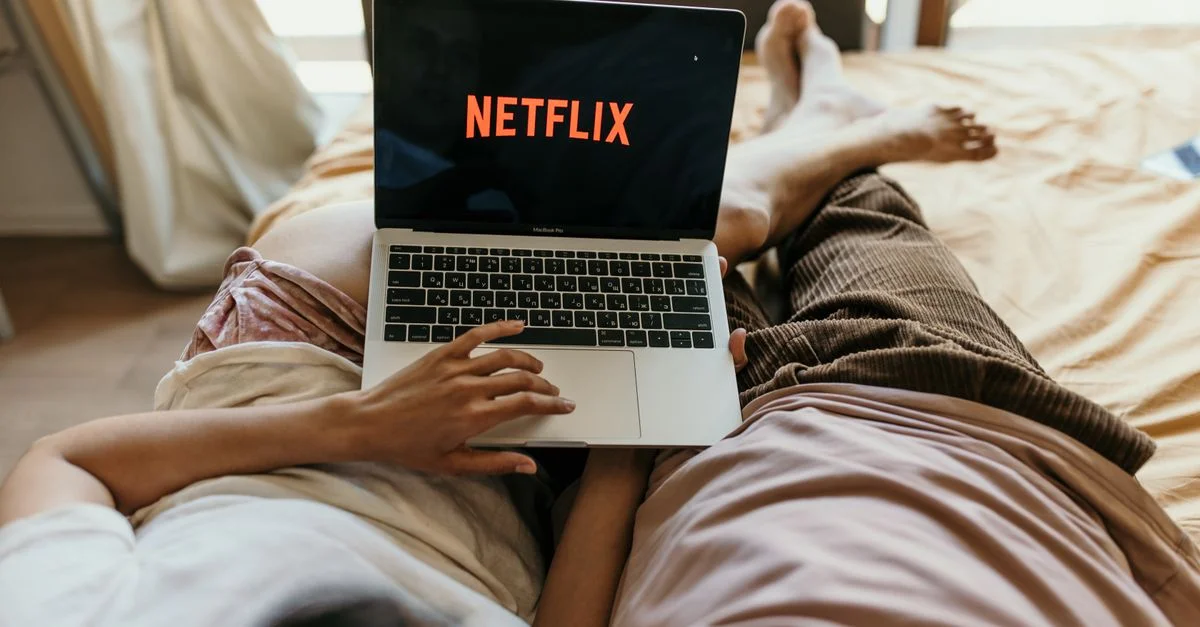 Una pareja acostada cómodamente en la cama viendo Netflix en un portátil, disfrutando de tiempo libre juntos en casa.