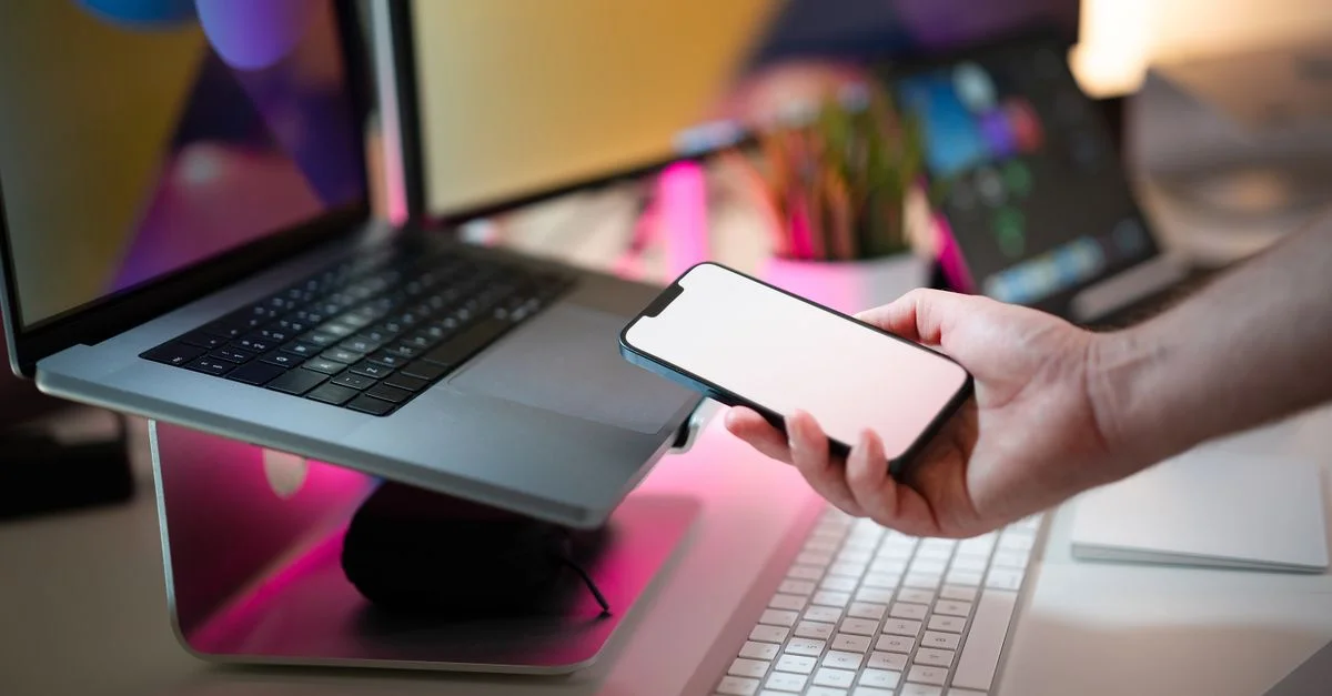 Primer plano de una mano sosteniendo un smartphone con pantalla en blanco junto a un portátil en una oficina moderna.