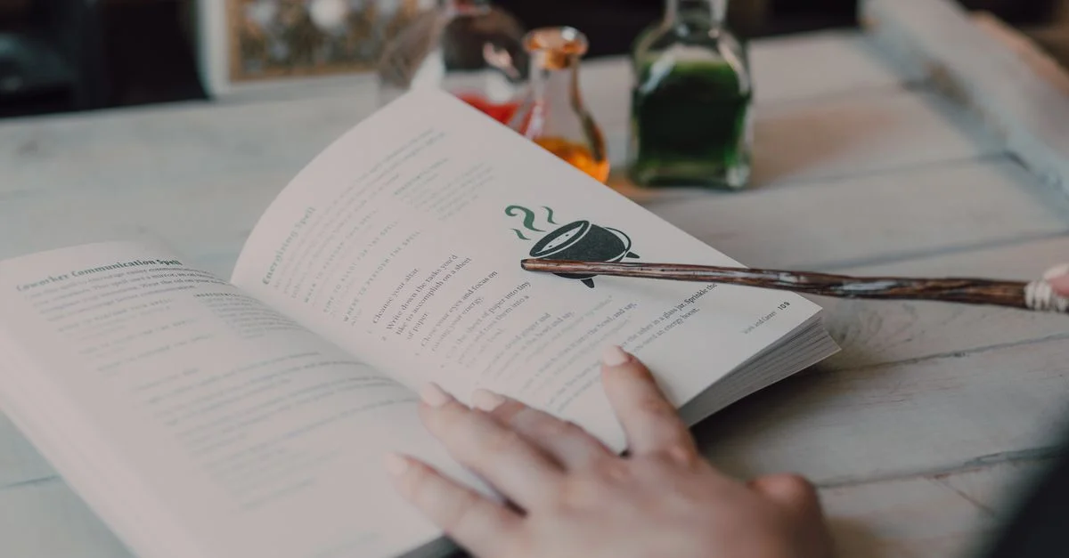 Una mano sosteniendo una varita señala un libro de hechizos con pociones mágicas de fondo.
