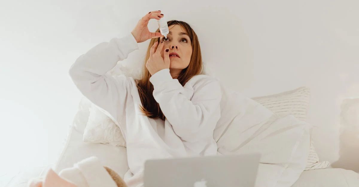 Una mujer aplicando gotas para los ojos mientras descansa en la cama con una laptop, simbolizando relajación y autocuidado en casa.