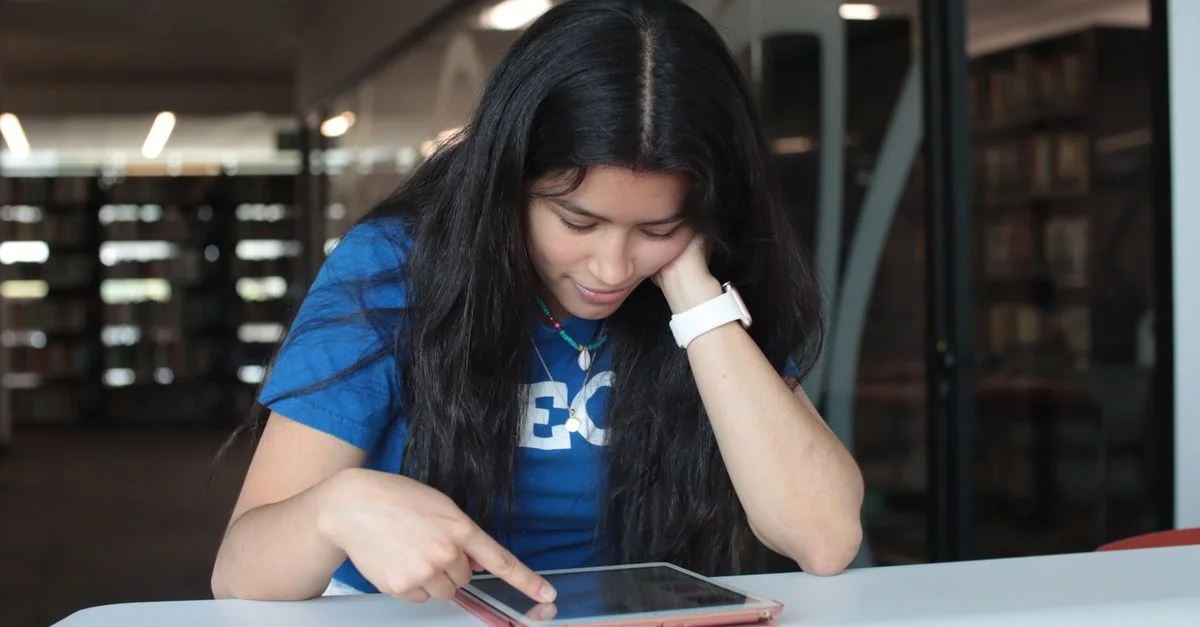 Una joven mujer con una camiseta azul está usando una tableta en una mesa de biblioteca. Enfocada y comprometida con su tarea.