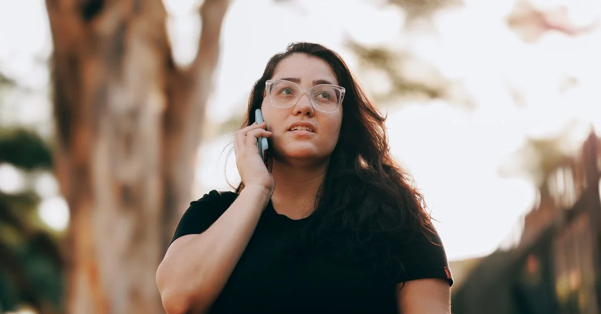 Joven mujer con gafas en camiseta negra hablando por teléfono móvil al aire libre.