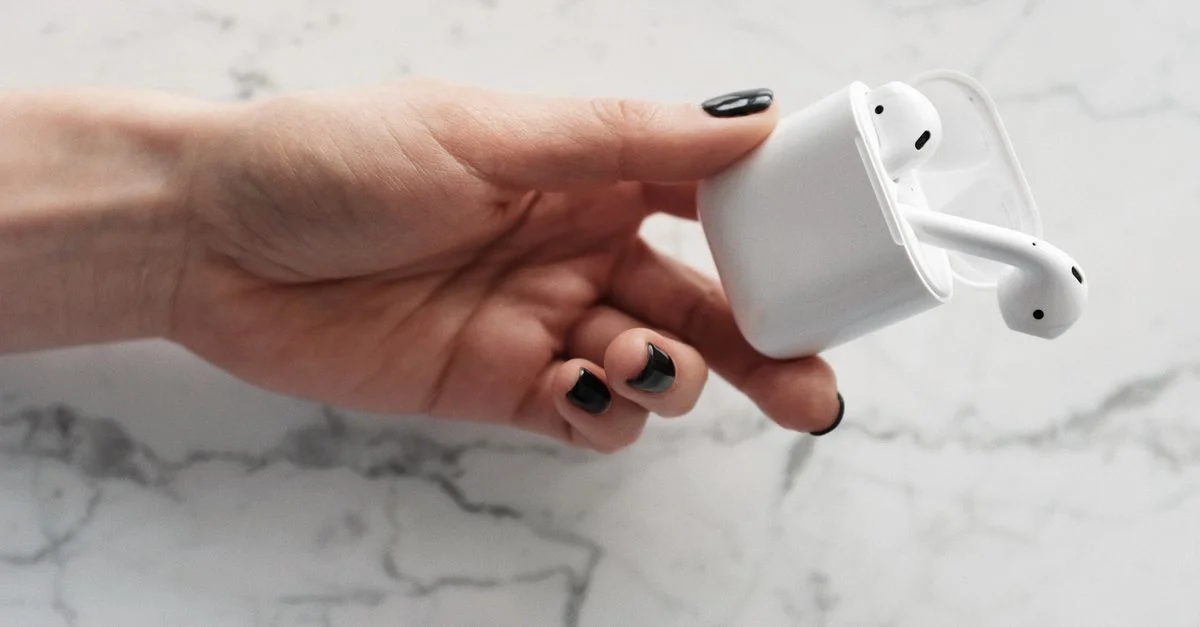 Mano con uñas negras sosteniendo auriculares inalámbricos sobre un fondo de mármol.