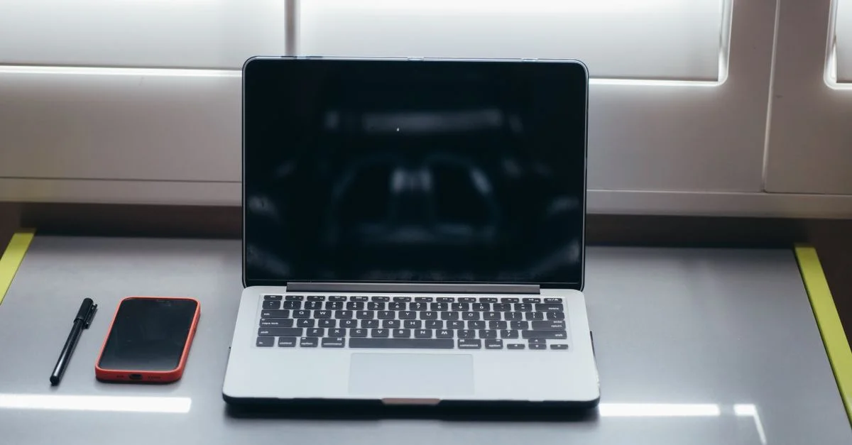 Una configuración de escritorio minimalista que presenta un portátil, un smartphone y un bolígrafo bajo una suave luz de ventana.