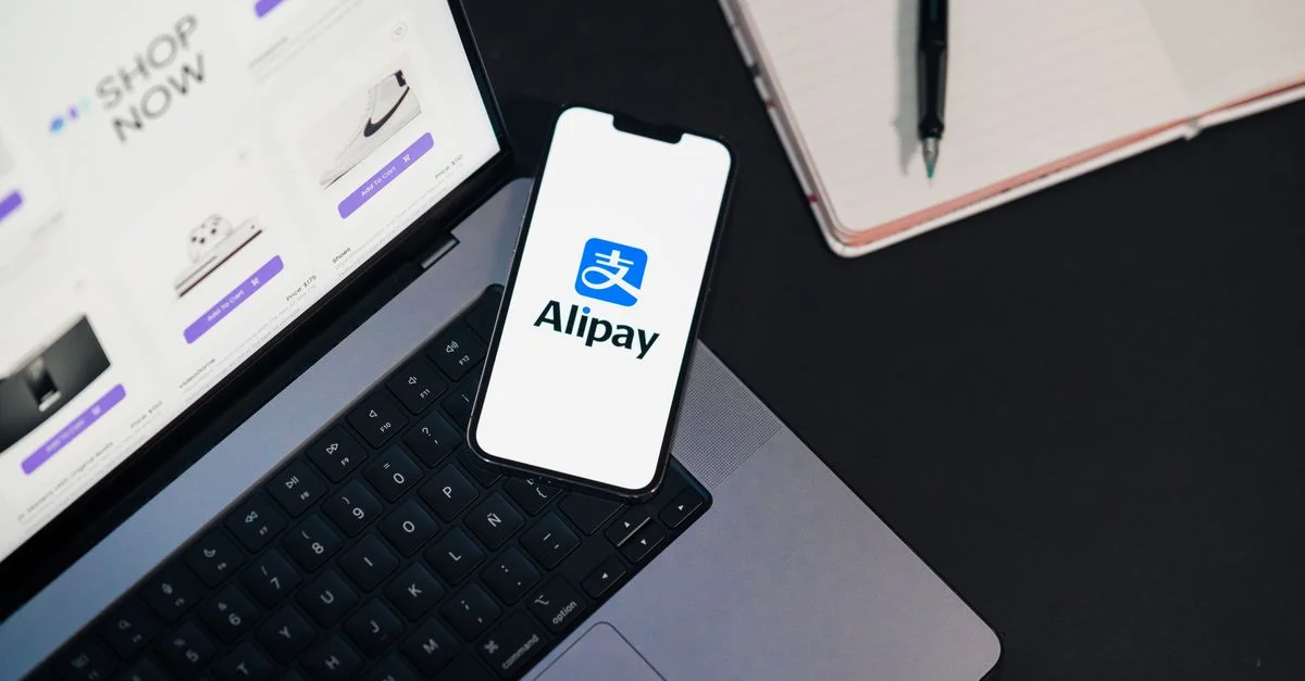 Teléfono inteligente que muestra la aplicación Alipay en una computadora portátil abierta con un sitio de compras en línea.