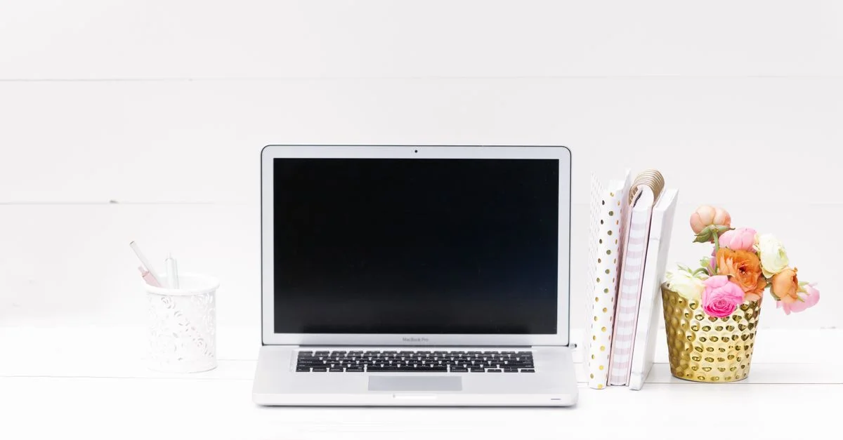 Espacio de trabajo minimalista con una computadora portátil, cuadernos y una vibrante decoración floral sobre una superficie blanca.