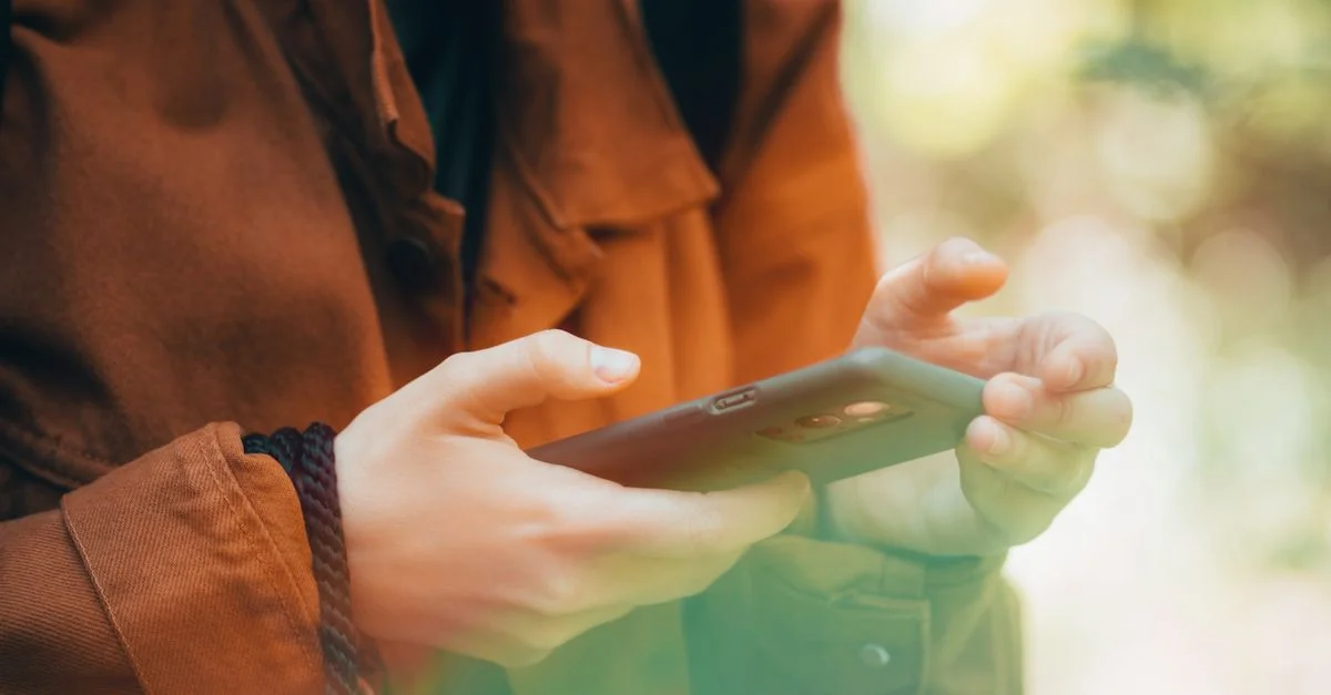 Primer plano de manos usando un teléfono inteligente al aire libre con un ambiente otoñal, mostrando tecnología y naturaleza.