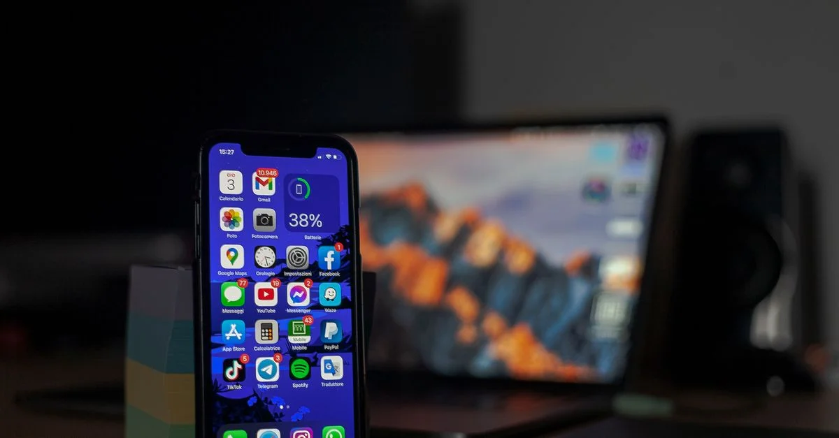 Smartphone mostrando varias aplicaciones en enfoque con un portátil desenfocado de fondo.