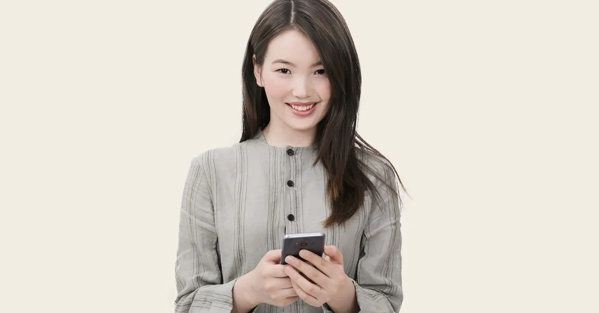 Mujer sonriente con cabello largo usando su smartphone, vestida con un atuendo casual contra un fondo simple.