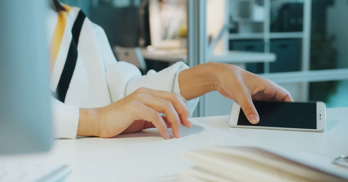 Primer plano de la mano de una mujer usando un teléfono inteligente en un escritorio en un entorno de oficina moderno.