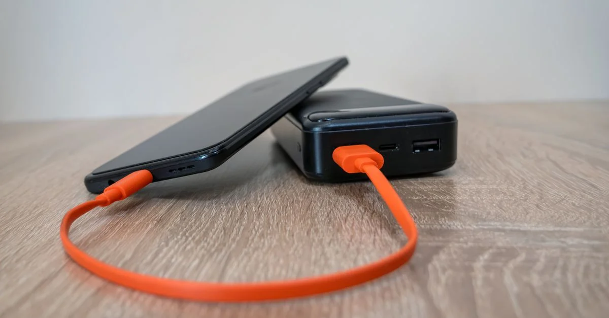 Un teléfono inteligente cargándose mediante un cable naranja conectado a una batería externa sobre una superficie de madera.