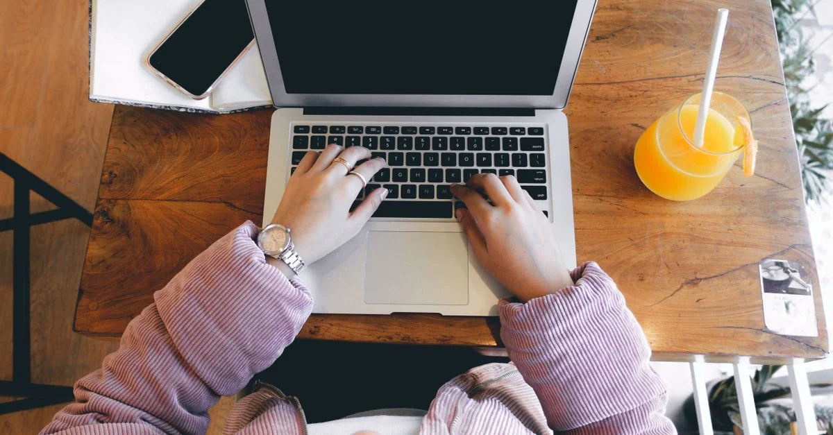 Vista en ángulo alto de una mujer usando un laptop en una mesa de madera con jugo de naranja y un smartphone.
