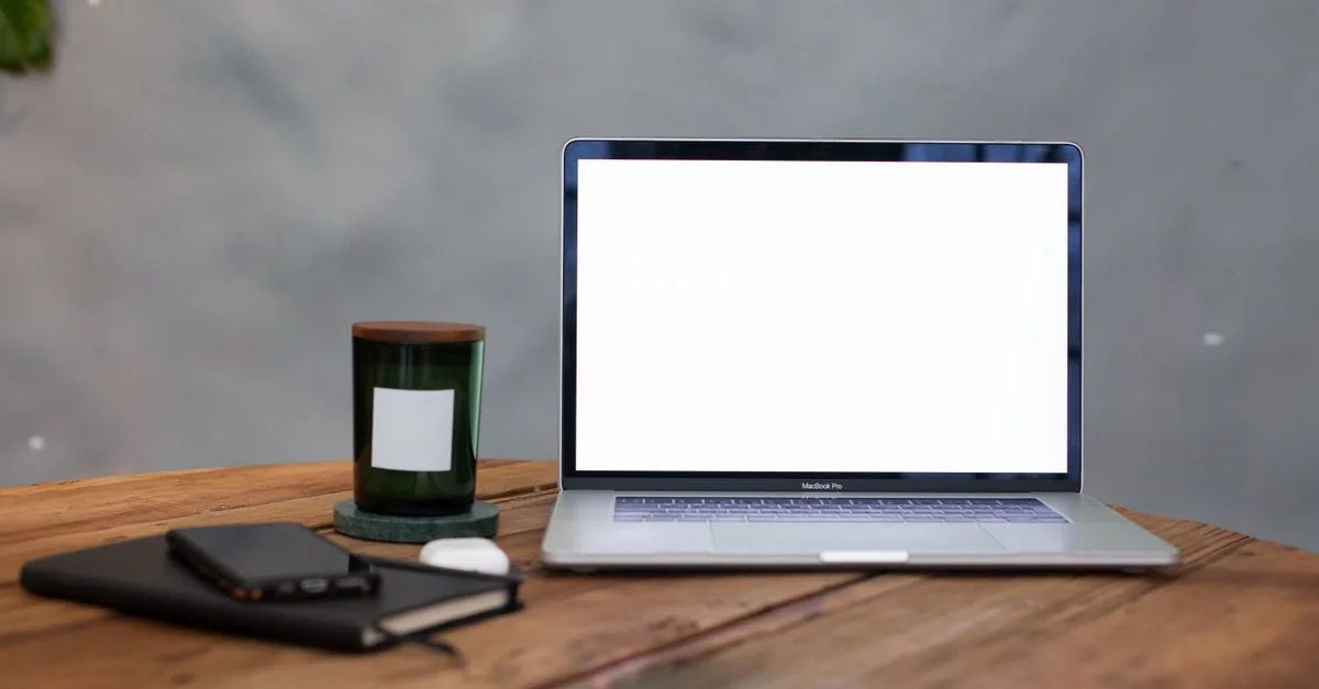 Un espacio de trabajo moderno con una pantalla de portátil en blanco, cuaderno y vela sobre una mesa de madera.