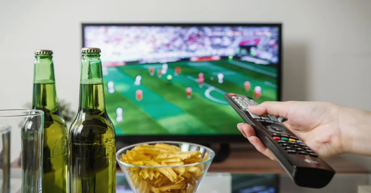 Una persona sosteniendo un control remoto mientras mira fútbol en la TV con cerveza y aperitivos en la mesa.