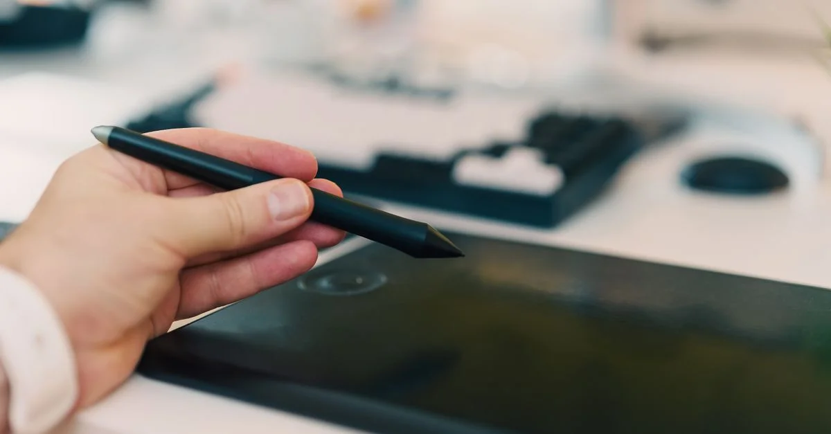 Primer plano de una mano que utiliza un lápiz óptico en una tableta digital en un entorno de oficina.